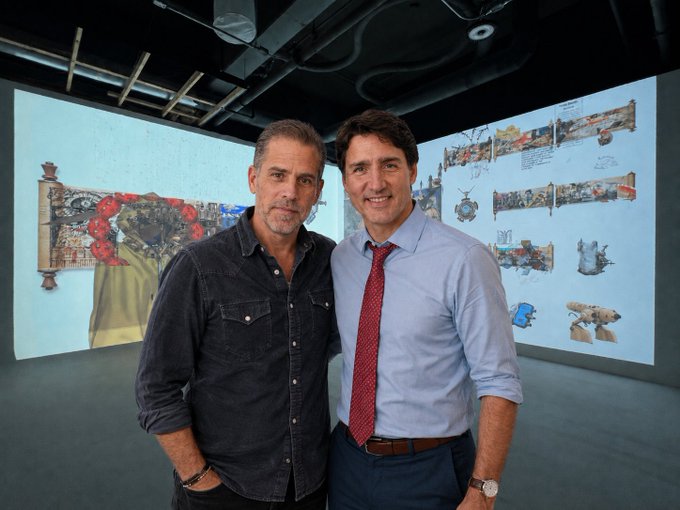 Two men stand together in a gallery space with projected art behind them; the man on the right resembles Canadian Prime Minister Justin Trudeau.