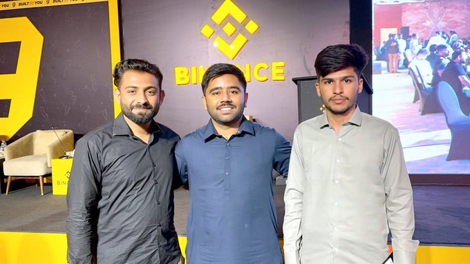 Three men pose together at a Binance event in Islamabad, standing in front of a yellow Binance-branded backdrop with the company's logo visible behind them.