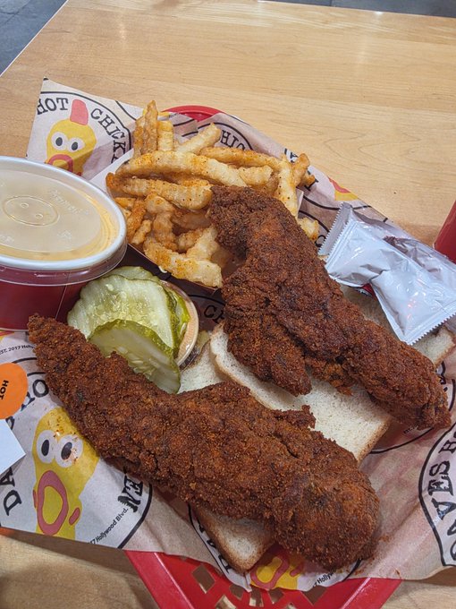 Overhead photo of a fast-food meal featuring two pieces of hot fried chicken on white bread with pickle slices, seasoned crinkle-cut fries, and a side of sauce in a paper-lined red tray.
