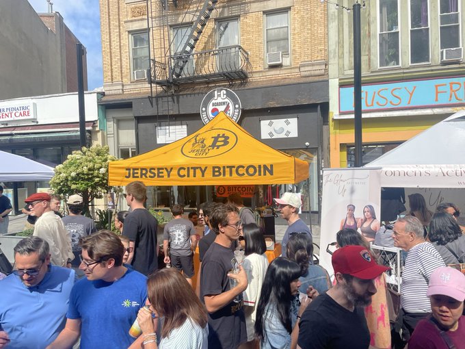 A bustling street fair features a bright orange tent advertising "JERSEY CITY BITCOIN" with a Bitcoin logo, set against urban storefronts.