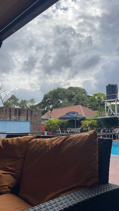 An outdoor patio or poolside view featuring orange cushions on wicker furniture in the foreground, with a swimming pool, umbrellas, a building with a red roof, and trees under a cloudy sky in the background.