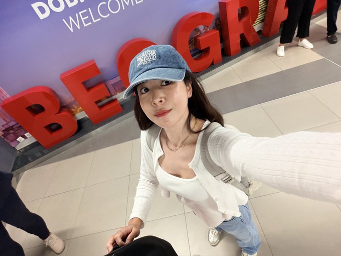 A woman wearing a denim cap and white cardigan takes a selfie in front of a display that reads "BELGRADE" with a "DOLLAR TREE WELCOME" sign.