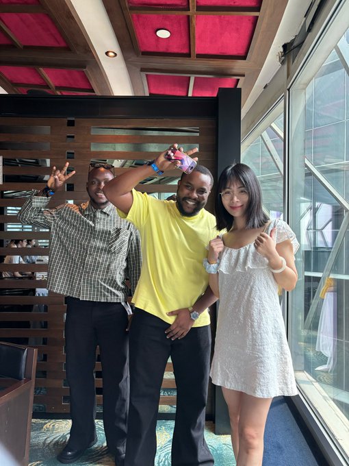 Three people pose smiling and gesturing toward the lens in a restaurant setting with a red-lit ceiling and large windows. The post celebrates a meeting with African supporters of the Axis project, suggesting the two men are community members (@Farmercist and @NDIDI_GRAM) joining in person after early contributions to building on Base.