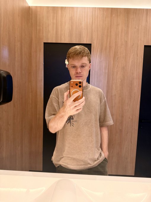 The image features a man taking a selfie with his cell phone, capturing himself in front of a mirror. He is wearing a brown t-shirt and has his hands in his pockets while holding the phone up to take the picture. The background includes a sink and a door, suggesting that he might be in a bathroom or a similar room. The post caption reads "Me waiting $poly", which could possibly be a reference to an event or gathering where people are expected to arrive soon.