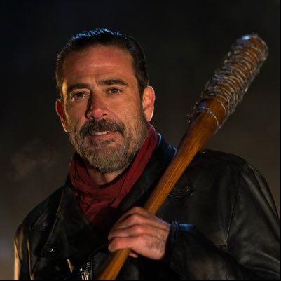 A man with a beard, wearing a black leather jacket and red scarf, holds a wooden baseball bat wrapped in barbed wire.