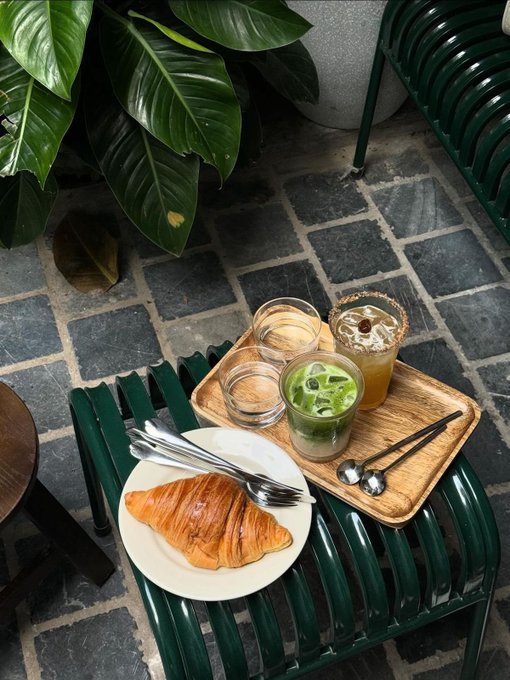 A wooden serving tray on a green slatted chair holds a croissant on a plate and three drinks, set on a tiled floor next to a leafy potted plant.