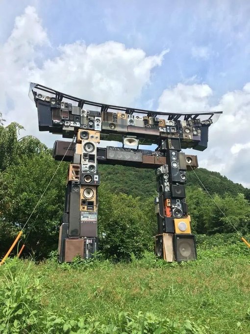 Large outdoor sculpture of a Japanese torii gate constructed entirely from stacked vintage speakers and audio equipment, set against a green field and cloudy sky.