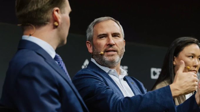 The image features three people, two men and one woman, all dressed in suits and wearing headphones around their necks. They are gathered together, with one of them giving a thumbs up gesture. The background appears to be a dark room or stage setting, which could suggest that they might be at an event or conference related to cryptocurrency or blockchain technology.