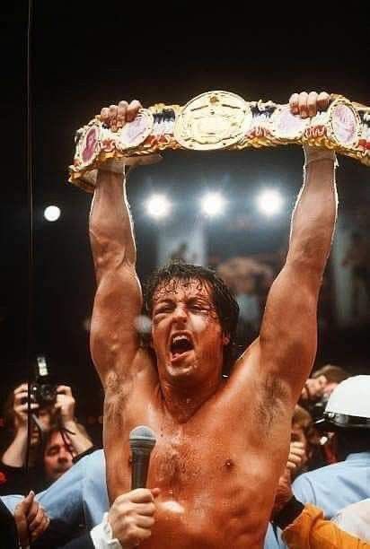A muscular, shirtless professional wrestler triumphantly raises a championship belt above his head while holding a microphone, surrounded by a cheering crowd in an arena.