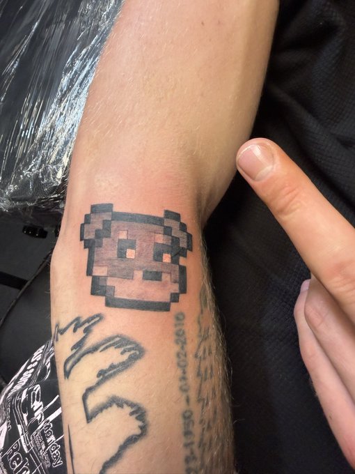 A freshly inked pixelated dog tattoo sits on a forearm, accompanied by the numbers "1956-02-2810," alongside other tattoos and a pointing finger—capturing a new addition to the wearer's collection as they showcase pride in the Doginal Dogs community.