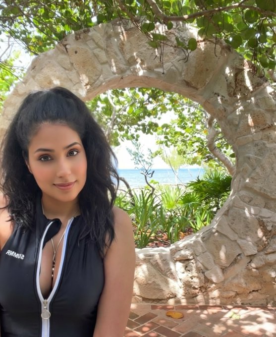 A woman poses in front of a large circular stone archway with tropical foliage and ocean visible through the opening, wearing a black SUNUV2 branded rashguard. The serene beachside morning setting complements the "Good morning" greeting, suggesting a relaxing start to the day in a tropical destination.