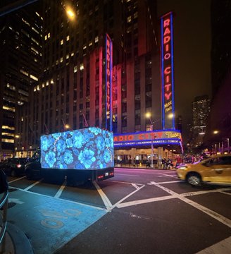 A nighttime street view of the Radio City Music Hall in New York City, featuring its iconic vertical neon sign and a bright LED display in the foreground.
