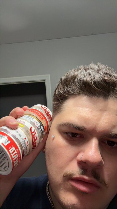 A man with short brown hair and facial piercings takes a close-up selfie while holding a can of Coors Light beer against his head.
