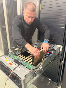 A man, reportedly the Czech central bank governor, works on a computer chassis containing eight graphics cards being repurposed into a mining rig, ironically showcasing hardware with a value negligible to the institution's substantial $170 billion balance sheet.