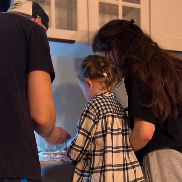 A candid family moment in a kitchen showing a man wearing a cap, a woman with long brown hair, and a young child with pigtails in a plaid shirt, all gathered together at what appears to be a counter.
