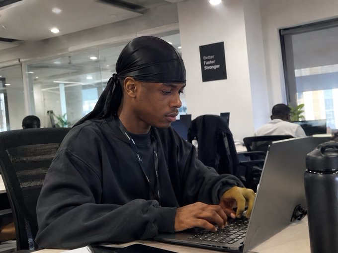 Photo of a man wearing a black durag and dark sweatshirt working on a laptop in an office setting with a motivational poster reading 'Better Faster Stronger' on the wall behind him.