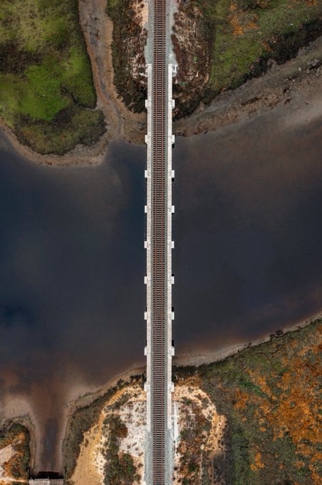 An aerial view captures a straight railway bridge spanning across a dark river or estuary, surrounded by marshy land with green vegetation and sandy banks.