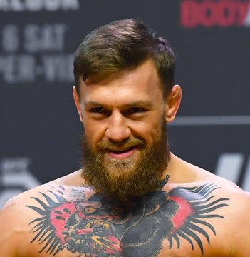 A photo of a heavily bearded, muscular man with extensive chest tattoos at what appears to be a UFC weigh-in or press event.