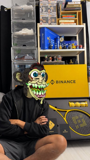 A person sitting cross-legged wearing a green monkey face filter, posing in front of a shelf filled with collectibles including Funko Pops and a yellow 'BINANCE' box and merchandise.