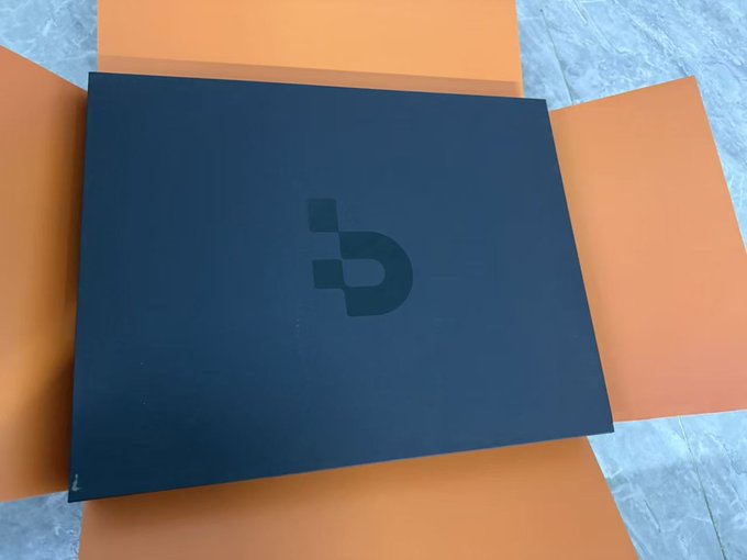 A matte black box with a stylized 'b' or 'd' logo sits inside an open brown cardboard shipping box.