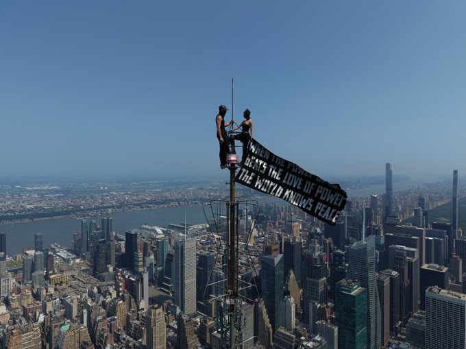 Two protesters perch atop a skyscraper antenna holding a banner reading "WHEN WILL THE BLACK BEATS THE LOVE OF POWER OF THE WORLD KNOWS PEACE" over the New York City skyline. The image contrasts the high-stakes, attention-grabbing spectacle of urban activism with the post's focus on an "Early Alpha" NFT collection building hype before its drop.