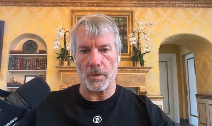 The image shows Michael Saylor, the executive chairman of MicroStrategy, appearing on a video broadcast while wearing a black t-shirt featuring the Bitcoin logo. The elegant room behind him includes a decorative Bitcoin plaque on a shelf, reinforcing the connection to the post about Strategy sharing an investor guide regarding a potential 93% drop in Bitcoin.