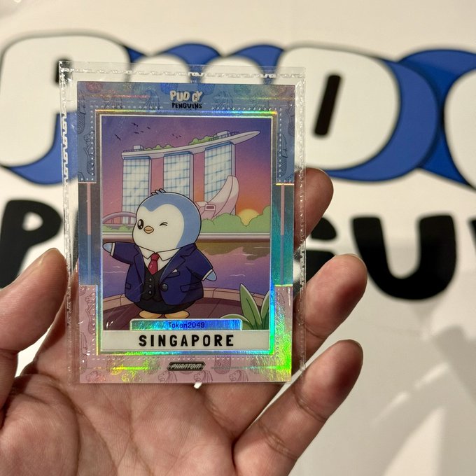 A hand holding a holographic "PUDGY PENGUINS" trading card featuring a penguin in a suit with the text "SINGAPORE" and "TOK2049".