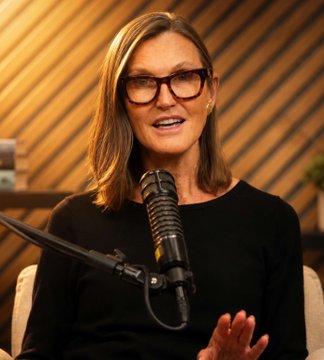 A woman with glasses and a black top speaks into a podcast microphone, with the post referencing Cathie Wood's bold Bitcoin price predictions of $730,000 base case and $1.5 million bull case by 2030, suggesting she is the speaker sharing her bullish crypto outlook during the interview.
