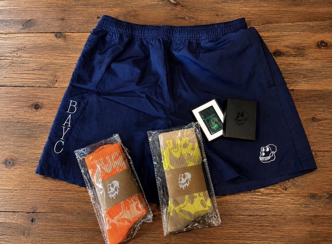 A flat-lay photograph on a wooden surface showing navy blue athletic shorts with "BAYC" lettering, two packages of orange and yellow socks with skull logos, and a black box, likely a promotional bundle.
