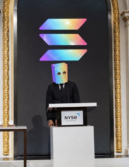 A person in a black suit stands at an NYSE (An ICE Exchange) podium wearing a holographic rainbow box over their head, with the Solana logo displayed prominently on the wall behind them.
