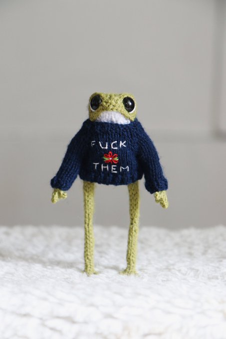 A small knitted amigurumi frog figure wearing a dark blue sweater with the text 'FUCK THEM' and a small flower design, standing on long green legs on a textured white surface.
