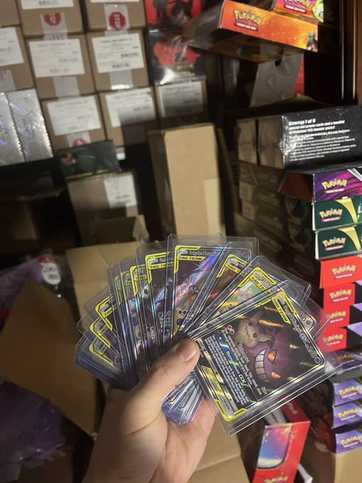 A hand fans out several Pokémon trading cards, including one labeled "GIGANTIC FALLING BOLT," in a room stacked with cardboard boxes and various Pokémon product boxes.