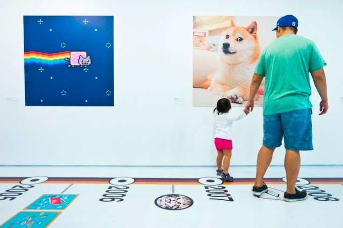 A man and young child view artworks at a gallery, including a Nyan Cat print and a large Shiba Inu dog image, while standing on a timeline floor display.