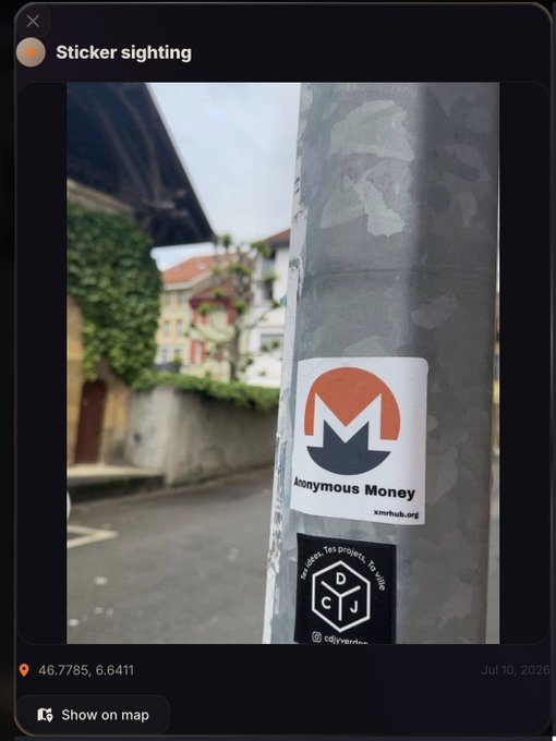 A Monero (XMR) "Anonymous Money" sticker from xmrhshub.org is affixed to a metal pole on a street, posted as a "Sticker sighting" on what appears to be XMRMap.org with coordinates 46.7785, 6.6411, illustrating the platform's new feature allowing users to geotag and upload crypto-related stickers spotted in the real world.