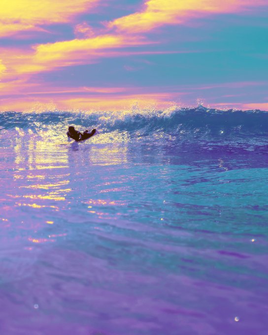 A serene ocean scene at sunset featuring pastel pink, purple, and blue colors, with a surfer riding a wave in the distance.