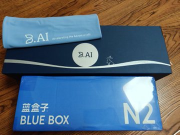An 'BAI - Accelerating the Advent of AGI' branded bag and a blue '蓝盒子 BLUE BOX N2' package are stacked on a wooden floor.
