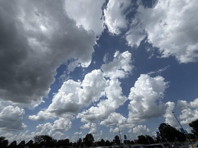 A dramatic sky photograph showing large, fluffy cumulus clouds against a deep blue sky, with trees, light poles, and parking lot structures visible along the bottom edge.
