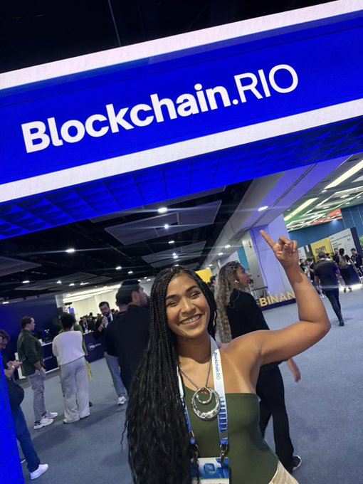 A smiling young woman with long dark braids points up at a large blue "Blockchain.RIO" sign while attending the event, wearing a lanyard and large pendant necklace.