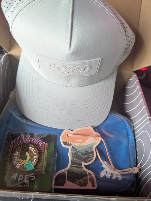 A light-colored trucker hat with 'BORED' embossed on the front patch, sitting in a box above a blue drawstring bag, an 'APES' welcome sticker, and an orange drawstring pouch.