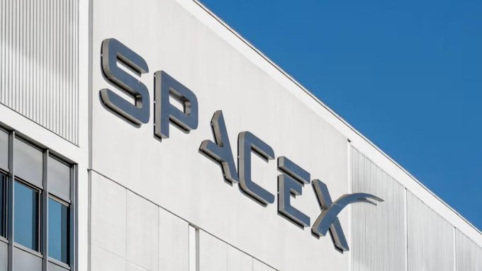 An exterior low-angle shot of a SPACEX building sign with silver metallic lettering mounted on a white paneled wall under a clear blue sky.
