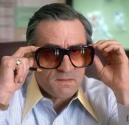 A man with grey hair puts on large, gradient-lensed sunglasses while wearing a striped shirt.