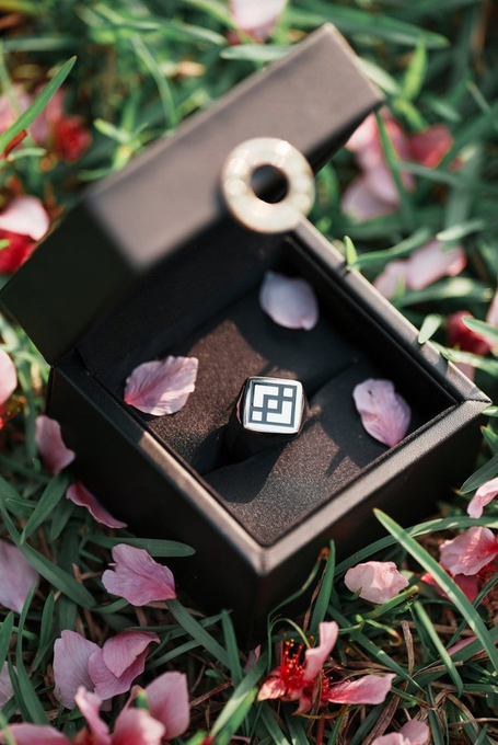A black jewelry box rests open on a bed of grass scattered with pink cherry blossom petals, revealing a silver ring with a geometric square-cut stone at its center. The petals surrounding the elegant ring box evoke the romantic sentiment of "Woke up and chose romance," transforming a simple morning moment into a scene of love and beauty.