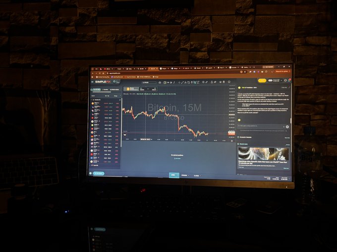 A computer monitor displaying a cryptocurrency trading chart showing Bitcoin's decline to 15M, set against a dark stone wall background.