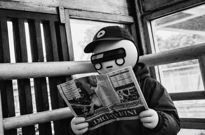 A black-and-white stylized photo of a character wearing a black cap, dark sunglasses, and a hoodie, sitting on a wooden bench while reading a newspaper titled "INDEPENDENT."