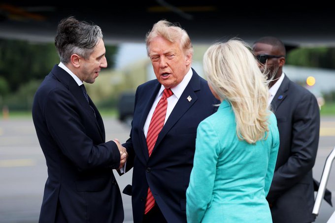 A photograph showing a man in a dark suit shaking hands with Donald Trump, who is wearing a navy blue suit and red striped tie, while a blonde woman in a turquoise blazer stands nearby and another man with sunglasses stands in the background.