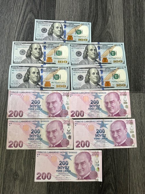 Five $100 USD bills are displayed above five 200 Turkish Lira (Türk Lirası) banknotes on a wooden surface, prompting a comparison between the two currencies' values. The image directly supports the question by visually presenting both options side-by-side for the viewer to evaluate.