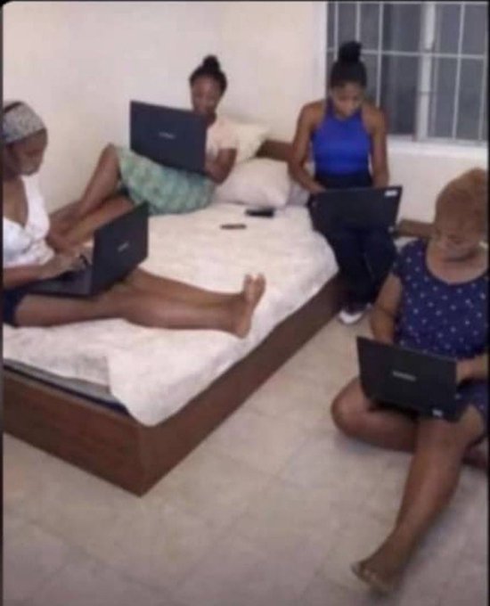 Four young women sit on and around a bed, each focused intently on their own laptop computers, seemingly absorbed in their individual screens rather than interacting with one another.