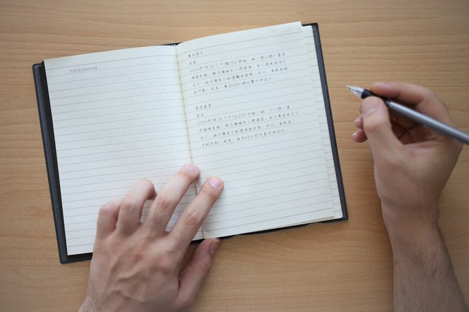 An overhead view of hands writing in a lined notebook with a silver pen, with Chinese text visible on the right page.