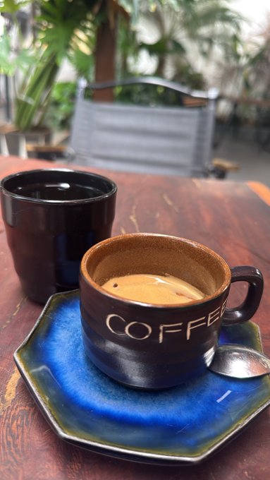 A small dark brown coffee cup labeled 'COFFEE' containing a shot of espresso sits on a blue ceramic saucer with a spoon, next to a larger dark cup, on a wooden table.