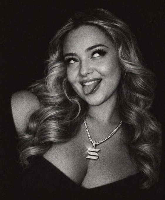 Black and white portrait of a smiling young woman with long wavy hair, wearing a necklace with a pendant, playfully sticking her tongue out.